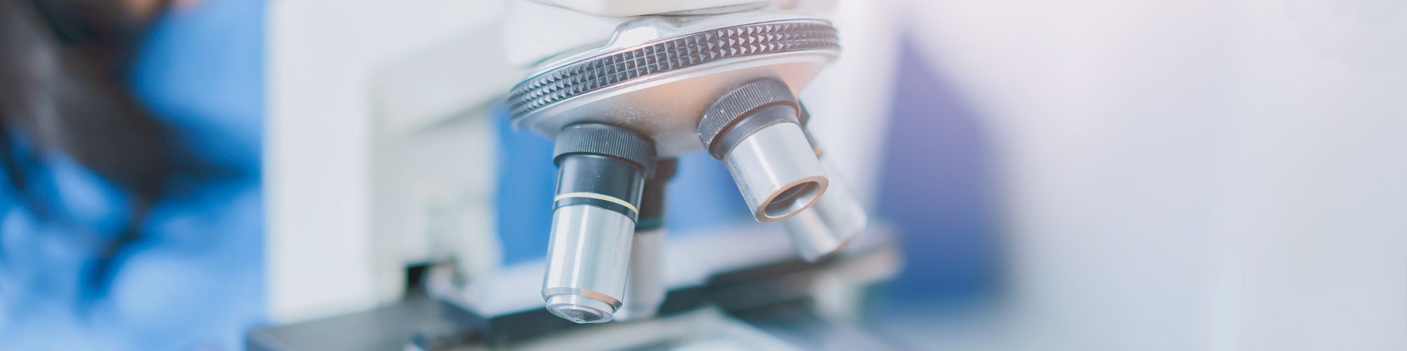 Close-up of a microscope, featuring two objective lenses and a rotating turret. The background is blurred, suggesting a laboratory environment. The image emphasizes the microscope's detailed components, highlighting its importance in scientific research.