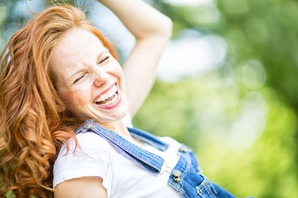 A woman with long, wavy red hair is joyfully laughing, her eyes closed and a huge smile on her face. She is wearing a white t-shirt under blue overalls, and her carefree expression suggests happiness and playfulness in a bright, outdoor setting.