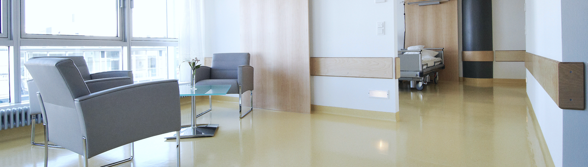 A modern hospital room featuring two gray armchairs and a glass table. The space is brightly lit with large windows allowing natural light. In the background, there is a hospital bed, creating a clean and open atmosphere. The flooring is smooth and yellow.