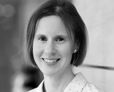 Das Bild zeigt eine lächelnde Frau mit schulterlangen, lockigen Haaren. Sie trägt ein helles Oberteil mit kleinen Punkten. Der Hintergrund ist verschwommen, wodurch der Fokus auf ihr freundliches Gesicht und Lächeln gelegt wird.