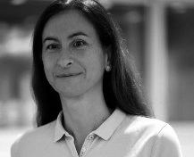 Eine Person mit langen, dunklen Haaren trägt ein helles Polohemd und schaut freundlich in die Kamera. Der Hintergrund ist unscharf und hell, was den Fokus auf die Person lenkt.