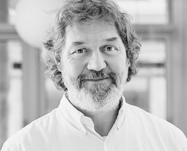 A middle-aged man with a friendly smile and a full beard is looking directly at the camera. He is wearing a white shirt and appears to be in an indoor setting with soft lighting. The image is presented in black and white.