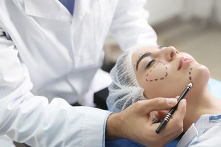 A medical professional in a white coat is marking a patient’s face with dotted lines to outline areas for a cosmetic procedure. The patient is lying on an examination table, wearing a head covering, while the clinician focuses on the markings.