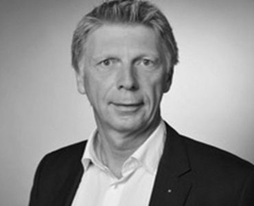 Porträt eines Mannes mit kurzen, grauen Haaren und einem seriösen Gesichtsausdruck. Er trägt ein schwarzes Jackett über einem weißen Hemd. Der Hintergrund ist neutral gehalten, um den Fokus auf die Person zu legen.