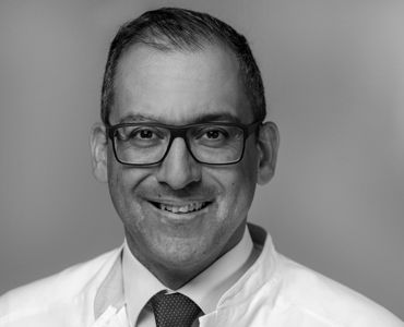 Ein lächelnder Mann trägt eine Brille und einen weißen Laborkittel. Der Hintergrund ist neutral gehalten, wodurch der Fokus auf seinem freundlichen Gesicht liegt.