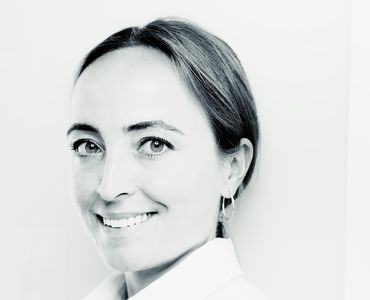 Porträt einer Frau mit langen, braunen Haaren und einem freundlichen Lächeln. Sie trägt eine weiße Bluse und hat Ohrringe an. Der Hintergrund ist hell und neutral. Ihre Augen strahlen Selbstbewusstsein und Freundlichkeit aus.