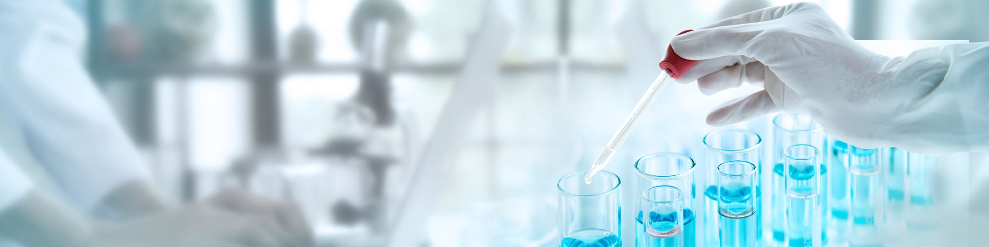 A laboratory scene featuring a gloved hand using a dropper to add a red liquid to a series of test tubes filled with blue liquid. The background includes blurred laboratory equipment and a person working at a desk, emphasizing a scientific research environment.
