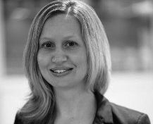 Porträt einer lächelnden Frau mit langen, hellen Haaren, die in einem professionellen Outfit gekleidet ist. Der Hintergrund ist unscharf. Die Frau wirkt freundlich und zugänglich.