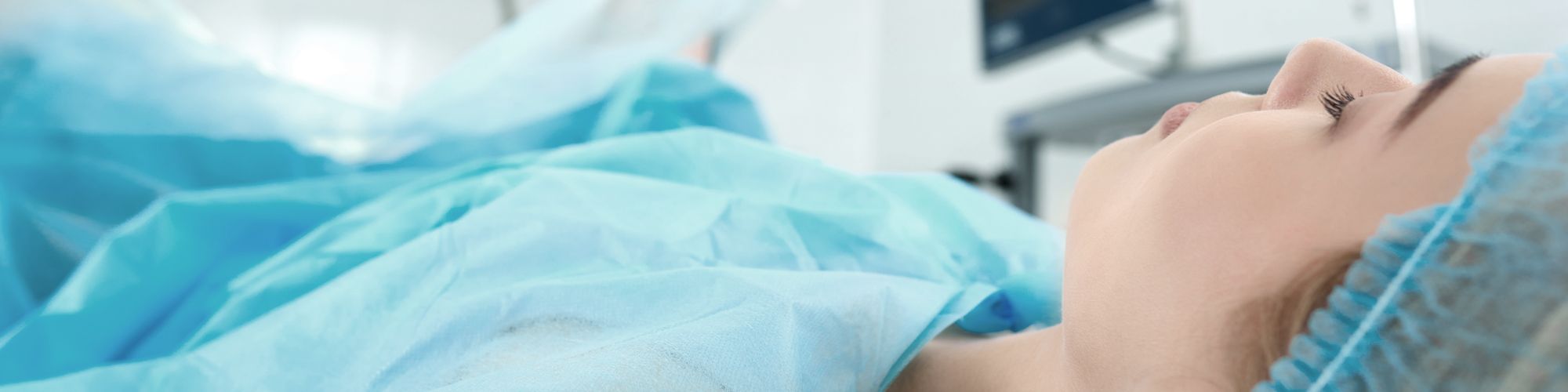 A patient lies on an operating table, covered with a light blue surgical drape. The scene suggests a medical environment, with a focus on the patient’s calm expression. Medical equipment is subtly visible in the background, indicating a surgical or clinical setting.