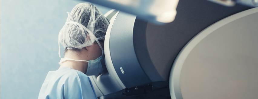 A healthcare professional in surgical attire and a face mask leans toward a high-tech medical machine, possibly preparing for a procedure. The environment suggests a clinical setting focused on patient care and advanced medical technology.