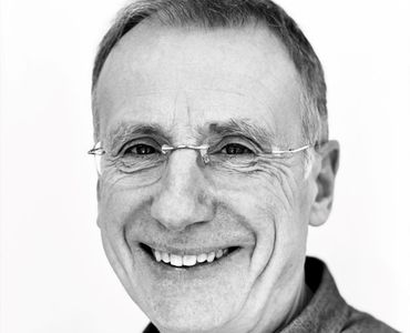 Porträt eines lächelnden älteren Mannes mit kurzen grauen Haaren und Brille. Der Hintergrund ist hell und neutral, wodurch der Fokus auf seinem freundlichen Gesicht liegt.