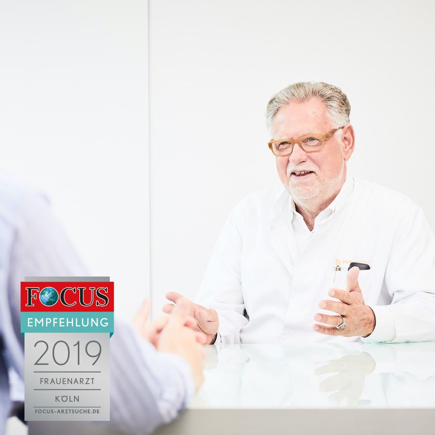 Ein lächelnder Arzt im weißen Kittel sitzt an einem Tisch und spricht mit einem Patienten. Der Raum ist hell und einfach gestaltet. Im Vordergrund sind Teile von einem anderen Menschen sichtbar, der den Arzt ansieht.