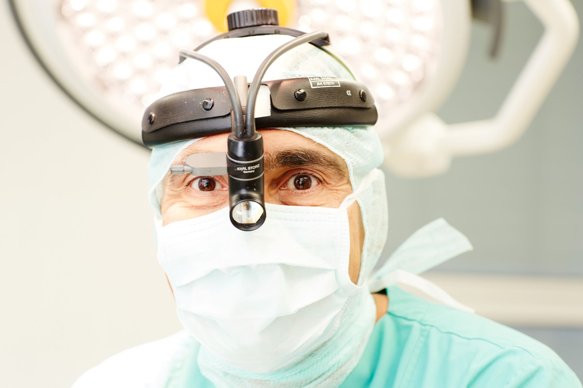 A surgeon in scrubs and a surgical mask is looking into the camera, wearing a headlamp for enhanced visibility. The setting suggests a medical environment, likely an operating room, emphasizing the professionalism and readiness for surgery.