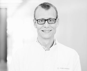 Das Bild zeigt einen lächelnden Mann in einem weißen Kittel mit einer Brille. Er steht in einem hellen Flur, der das Umfeld unscharf erscheinen lässt. Der Mann könnte ein Arzt oder eine Fachkraft im medizinischen Bereich sein.