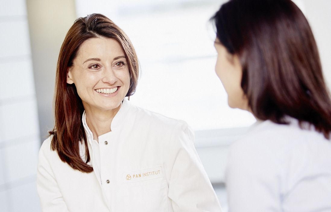 A female healthcare professional in a white coat is smiling and speaking to another woman seated across from her. The professional is holding a tablet and appears to be engaged in a friendly conversation, conveying a supportive and informative atmosphere.