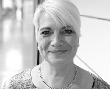 Porträt einer lächelnden Frau mit blonden, kurzen Haaren, die in einem hellen Raum steht. Sie trägt eine Kette und hat einen freundlichen Ausdruck. Der Hintergrund ist unscharf und zeigt einen modernen Innenraum.