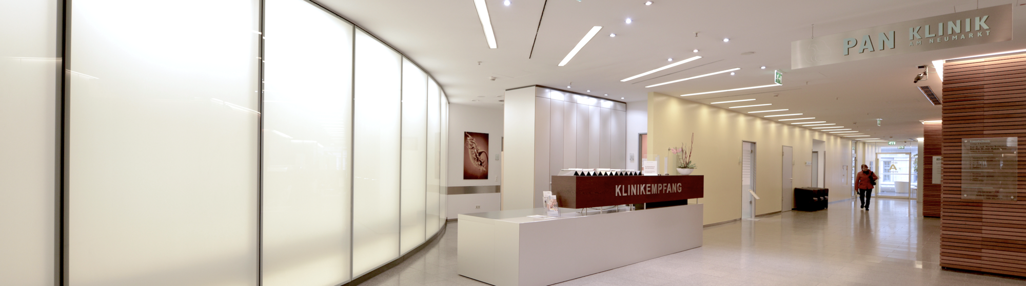 A modern, well-lit interior of a clinic features a reception desk with the words "KLINIK EMPFANG" prominently displayed. The space has smooth white walls and sleek design elements, with a person walking towards the entrance in the background.