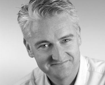 Ein lächelnder Mann mit kurzen, grauen Haaren trägt ein helles Hemd. Der Hintergrund ist neutral und hebt seine Gesichtszüge hervor.