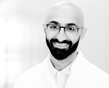 Ein lächelnder Mann mit Glatze und Brille trägt einen weißen Kittel. Der Hintergrund ist unscharf und hell. Der Ausdruck des Mannes vermittelt Freundlichkeit und Professionalität.