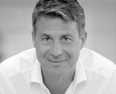 Ein lächelnder Mann mit grauem Haar und einem weißen Hemd schaut direkt in die Kamera. Der Hintergrund ist unscharf, wodurch der Fokus auf seinem Gesicht liegt.