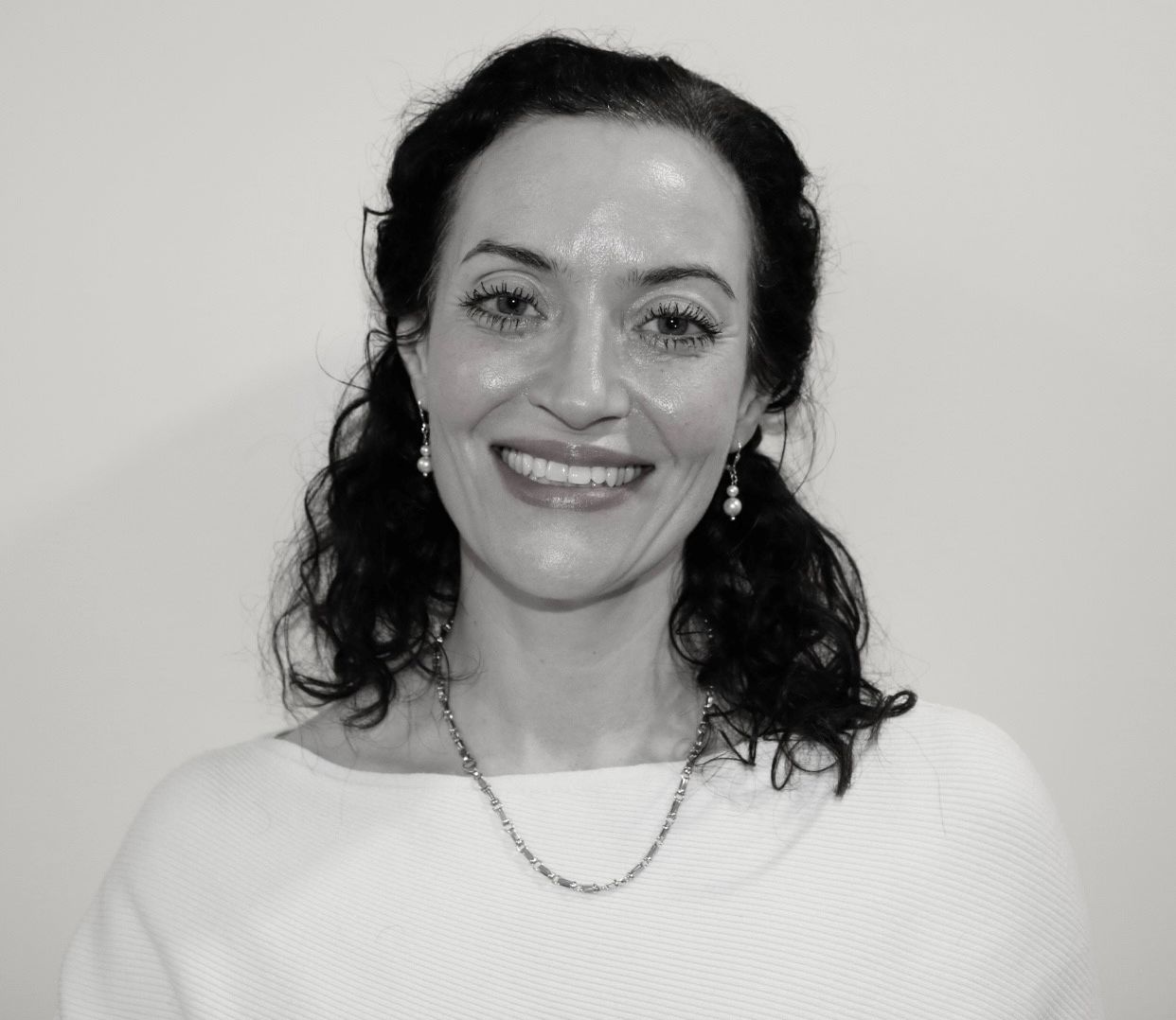 Ein Lächeln einer Frau mit lockigem, schwarzem Haar. Sie trägt ein helles Oberteil und eine Kette. Der Hintergrund ist einfach und hell. Die Atmosphäre wirkt freundlich und einladend.