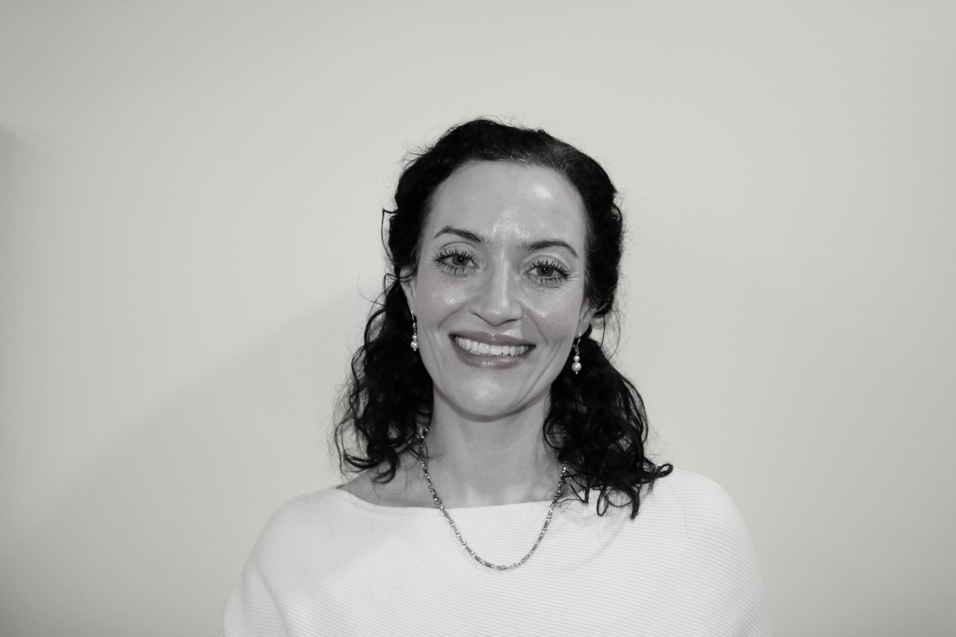 A woman with curly hair wears a white sweater and a necklace, smiling at the camera against a plain background. She has a friendly expression and is looking directly at the viewer.
