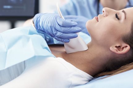 A woman lies on an examination table, receiving a medical procedure. A healthcare provider in blue gloves uses a device on her neck. The setting suggests a clinical environment focused on patient care.