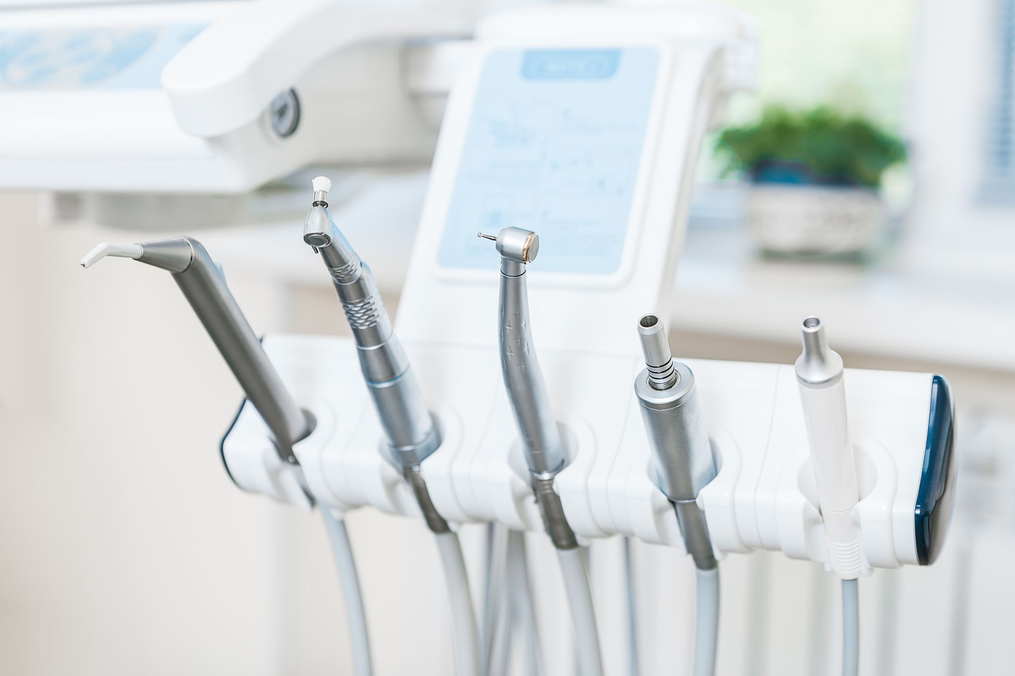 A close-up view of various dental instruments displayed in a holder, including a dental drill, scaler, and suction tool. The background suggests a clean, well-lit dental clinic environment.