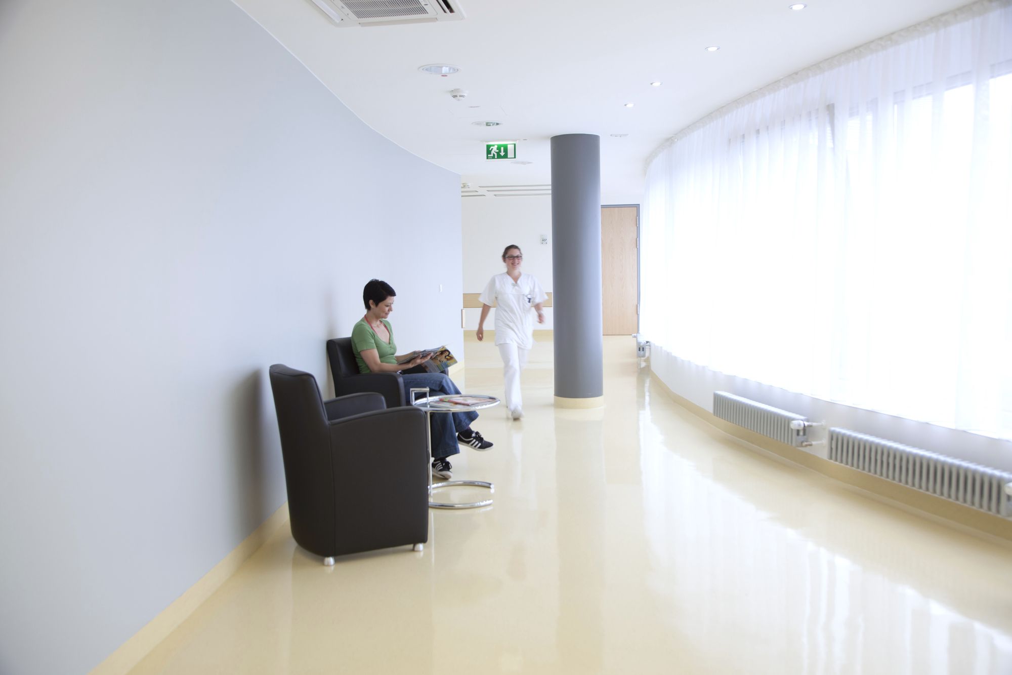 A well-lit corridor in a healthcare setting features a man sitting on a chair, reading. A healthcare professional in white attire walks toward him. The walls are smooth and light-colored, with natural light filtering through large windows.