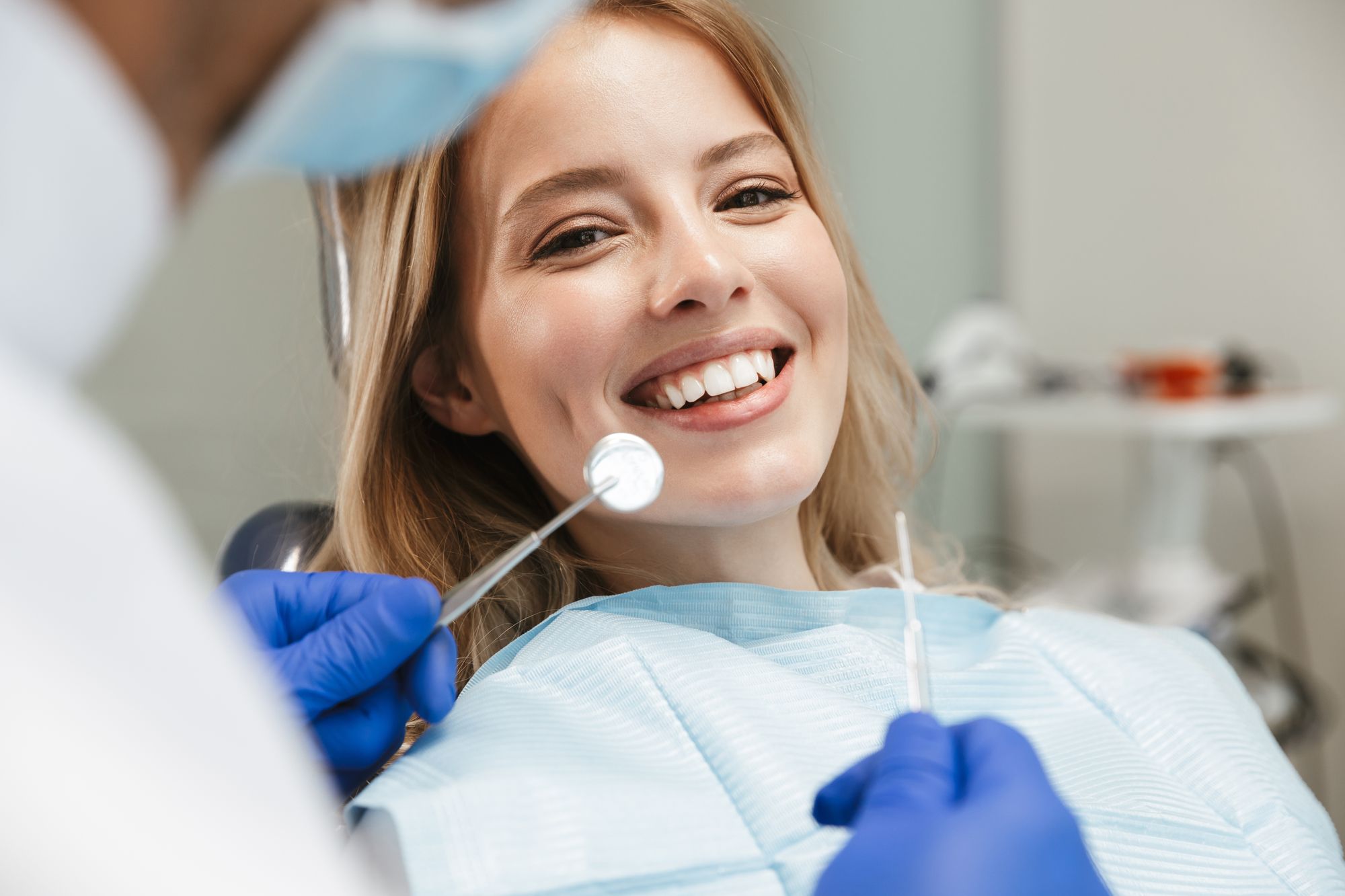 A smiling young woman sits in a dental chair, wearing a light blue bib. A dentist, partially visible, holds a dental mirror and tool, preparing for an examination. The setting is a modern dental office, emphasizing a positive dental care experience.