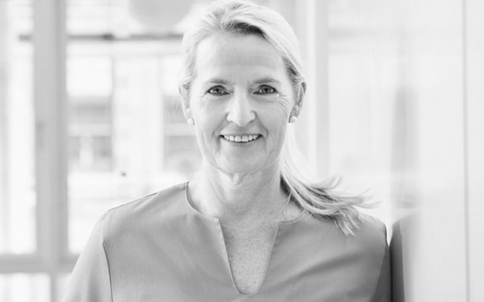 A smiling woman with long, blonde hair stands in a bright, modern setting. She wears a simple gray blouse and is positioned against a light-colored background, exuding a friendly and approachable demeanor.