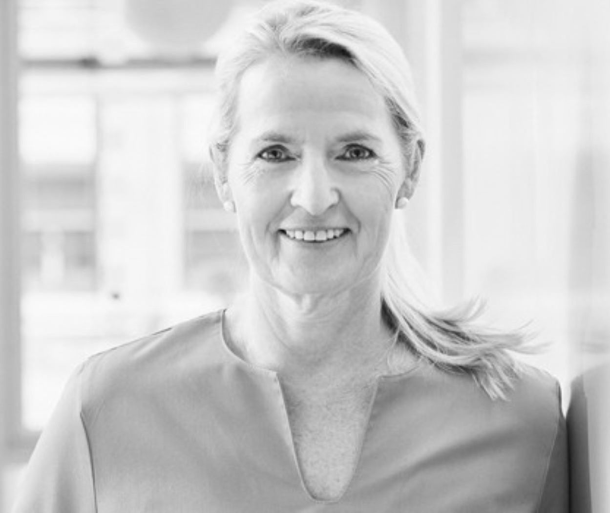 Eine blonde Frau mit schulterlangen Haaren lächelt freundlich. Sie trägt ein schlichtes, helles Oberteil und steht vor einer hellen Hintergrundkulisse. Die Atmosphäre wirkt freundlich und einladend.