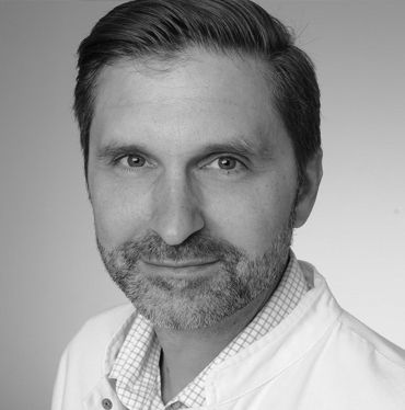 A middle-aged man with short, dark hair and a beard smiles at the camera. He is dressed in a white coat over a checkered shirt, set against a neutral background.