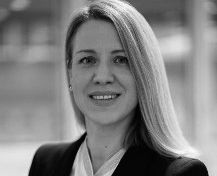 Porträt einer Person mit langen, blonden Haaren, die lächelt. Sie trägt einen schwarzen Blazer und ein weißes Oberteil. Der Hintergrund ist verschwommen und neutral.