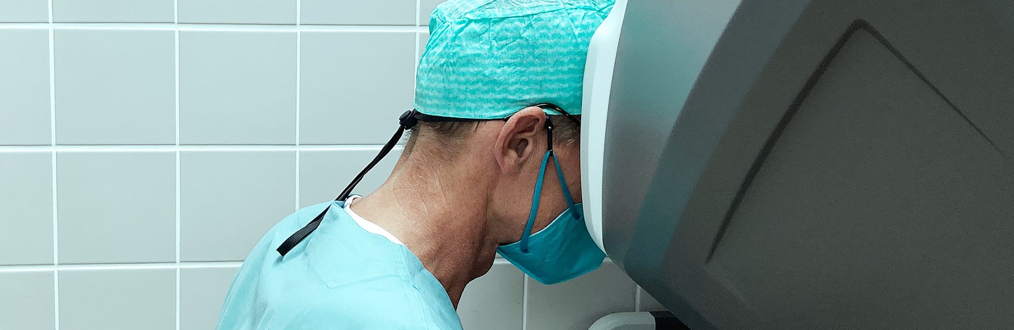 A surgeon in scrubs and a surgical mask is focused on operating a robotic surgical system. The environment is sterile, with light blue tile in the background. The surgeon’s attention is directed towards the equipment, indicating a precision medical procedure is taking place.